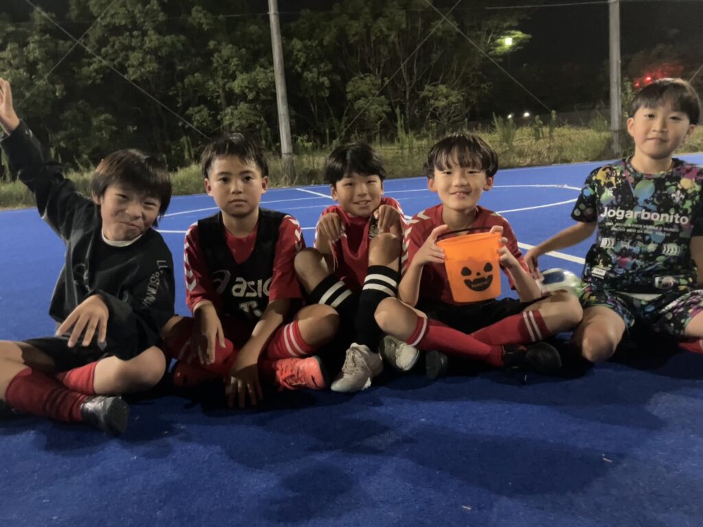 名古屋オーシャンズフットサルスクール桑名校のハロウィンイベントで、かぼちゃの容器を持って青いコートに座る5人のスクール生